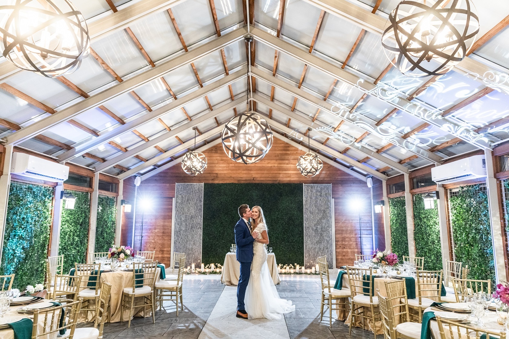 a wedding couple embracing in a large event space with a glass roof, wooden beams, and round tables set for a reception