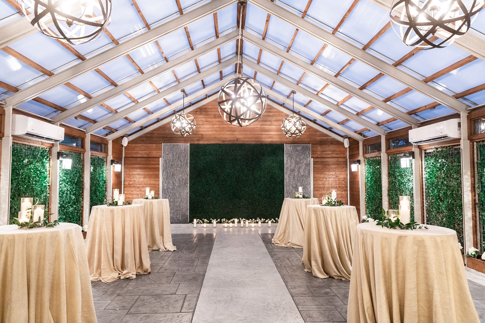 A elegantly decorated event space with round tables, candlelight, and a greenery wall under a skylight.